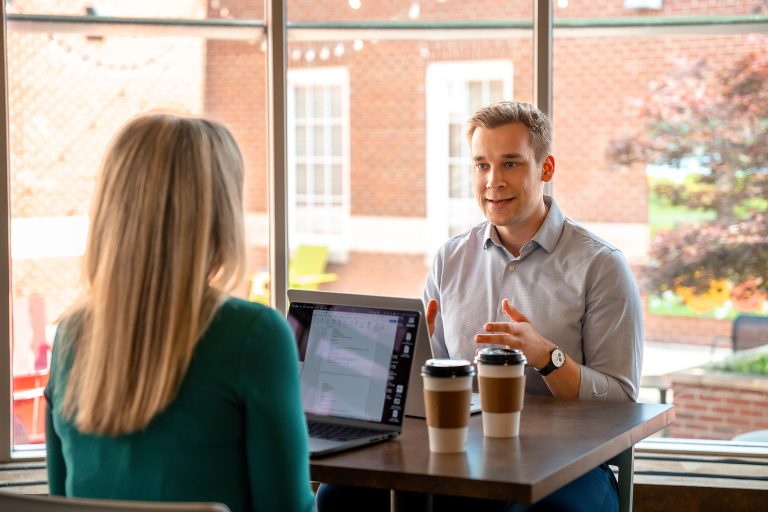 career coach lasse palomaki sits across a table from an alumni with a computer open and her back to the camera. Lasse is gesturing and giving advice