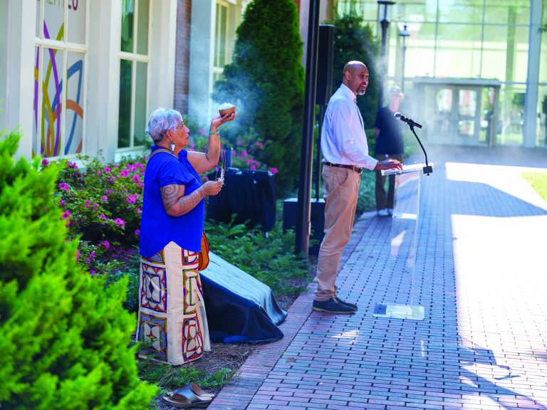 a member of the occaneechi band of the saponi nation holds a ceremonial bowl with smoke coming out of it. An Elon representative stands at a podium in front of the historical plaque before it is revealed