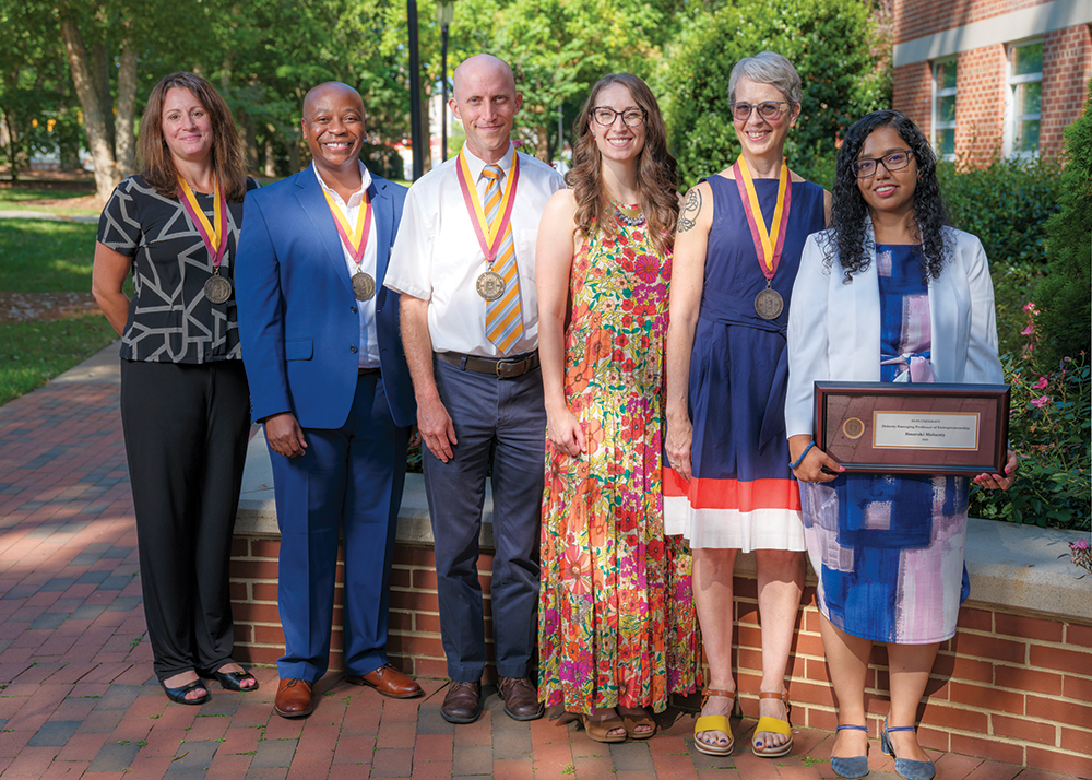 Six people stand in a line and smile at the camera while outside on a brick pathway underneath some trees with green leaves. All six people are Elon professors who were awarded endowed professorships in 2025