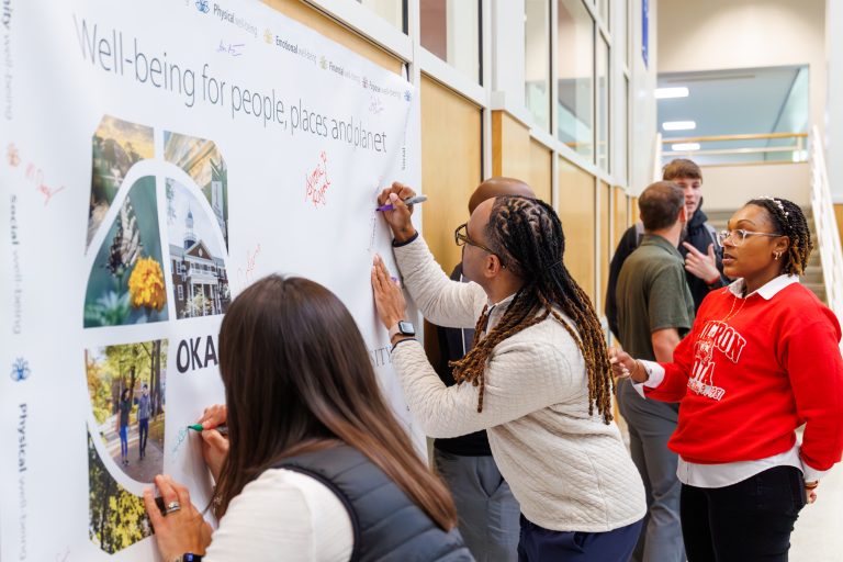 two people sign a poster hanging on a wall representing the Okanagan charter. One person watches and waits for their turn to sign.