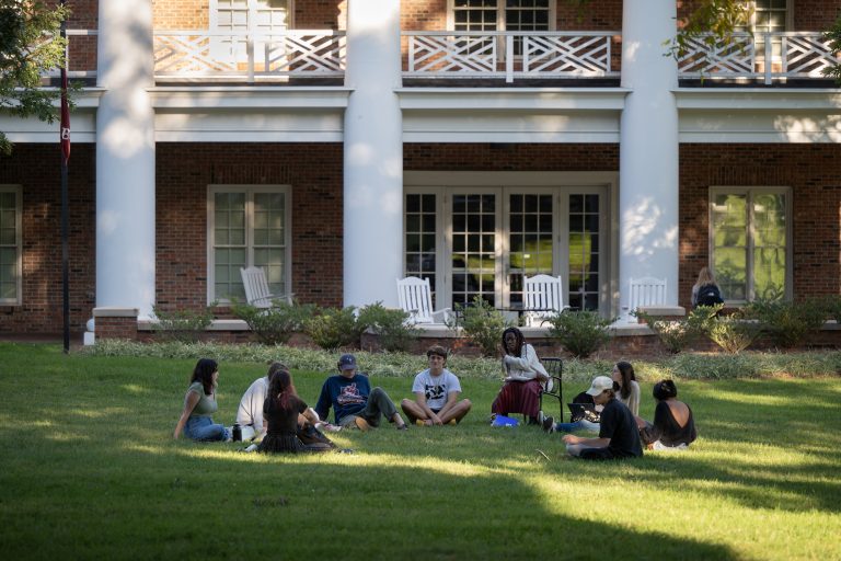 students and a teacher sit in a circleon the lawn outside of an academic building to have a discussion