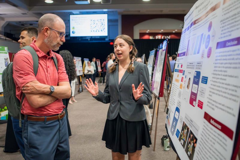 A person presents a research poster to another attendee at a conference, featuring various charts.