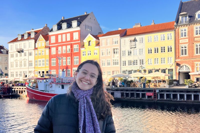Ellie Agulnek smiles beside Copenhagen’s Nyhavn canal, with a red boat and colorful waterfront buildings behind her.