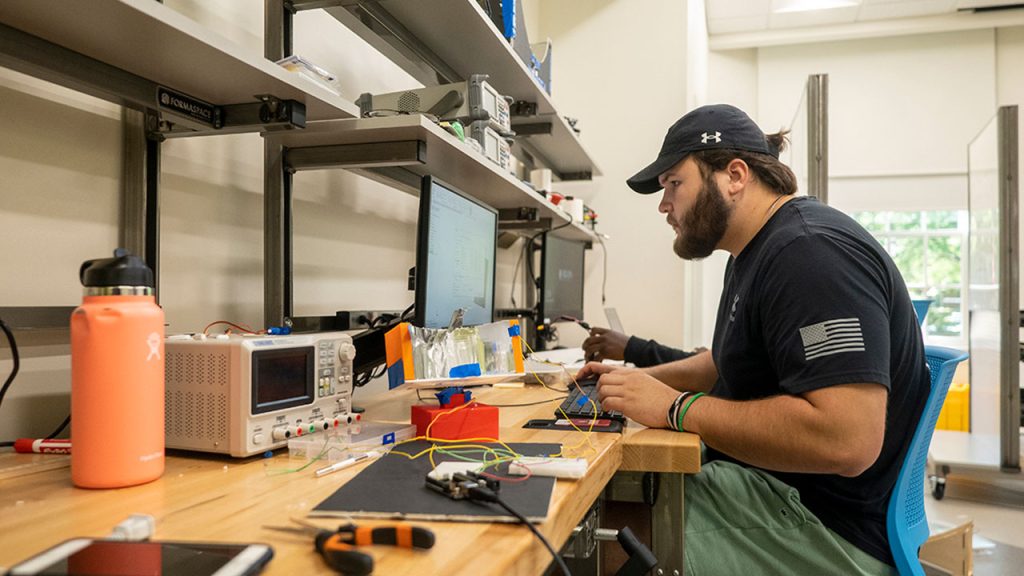 Student sits at work bench with computers, electrician's tools and various wires and boxes.