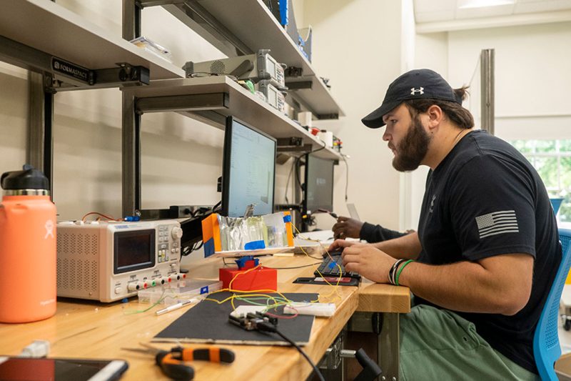 Student sits at work bench with computers, electrician's tools and various wires and boxes.