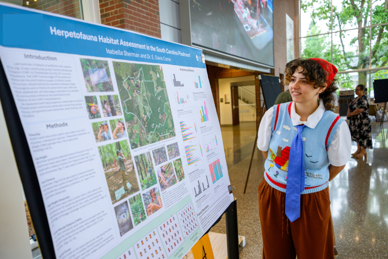 Student stands in front of project poster mapping habitats and comparing conditions across locations.