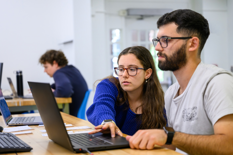Student leans in front of another to observe something on a laptop