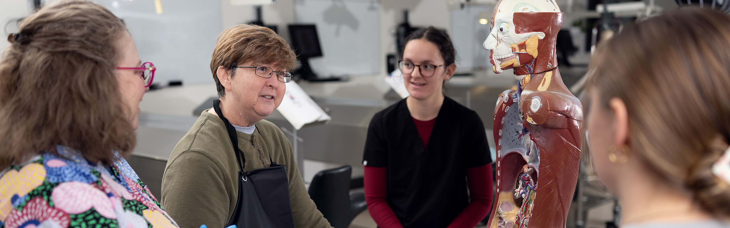 Faculty member speaks with students around an anatomical torso model in a lab.