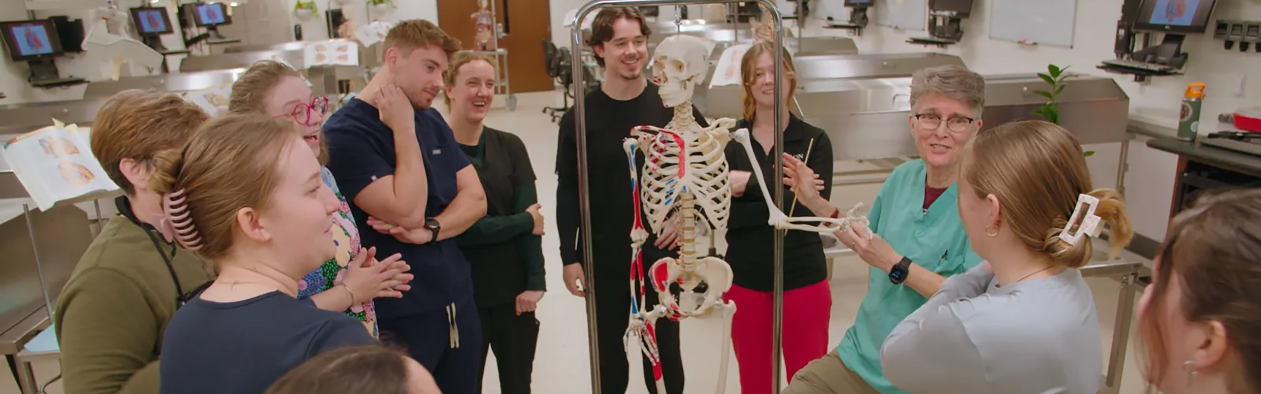 Faculty member leads a group of students studying a full skeleton model in an anatomy lab.