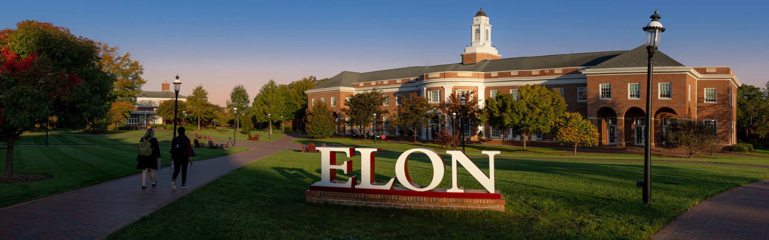 Human size letters spell ELON, and sit in the grass in front of a large brick building and students walking in the late afternoon sun