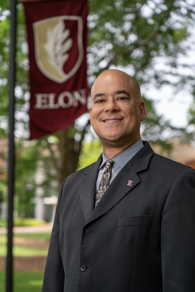 Kenn Gaither stands in a suit with an Elon flag flying behind him under the oaks.