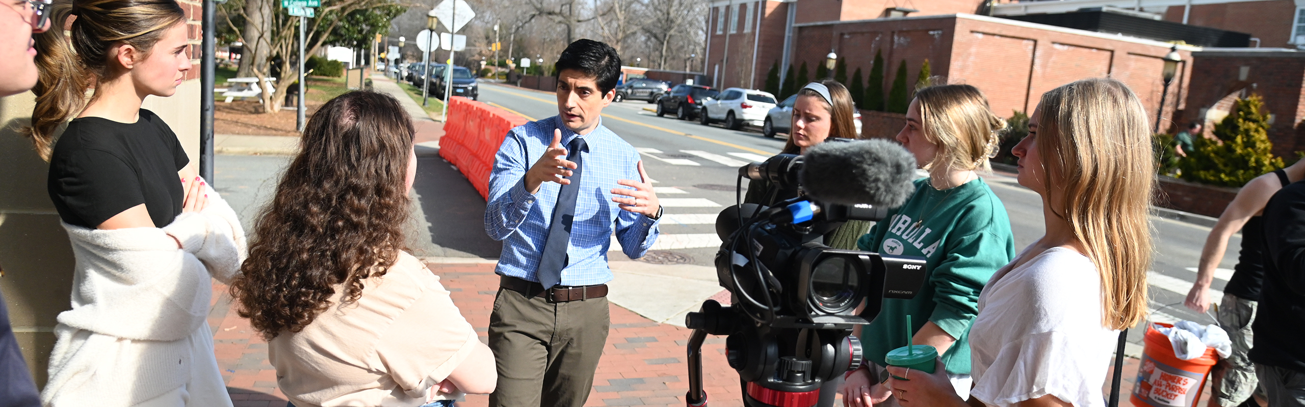 A group of students working toward a degree in journalism surround a professor as he talks on an Town of Elon street corner.