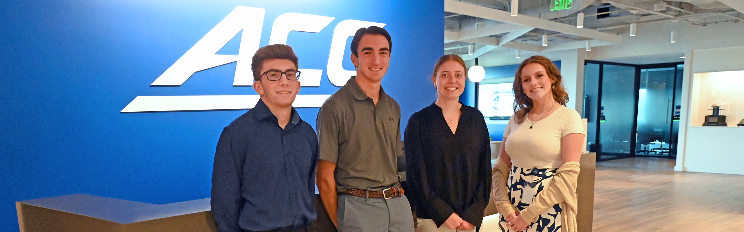 Four students stand in the entryway of the ACC offices in Charlotte in front of a blue wall.