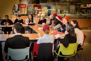 A group of students with red holiday themed hats gather around a table in Lakeside Dining hall to share a meal.  They are smiling and engaged in discussion. 