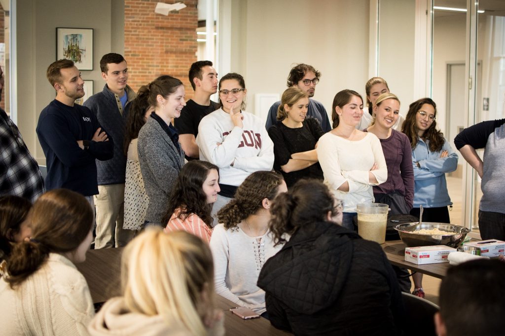 Students gather around tables that have been set up to prepare ingredients for a cooking class. The students are smiling and visibly excited about the experience.