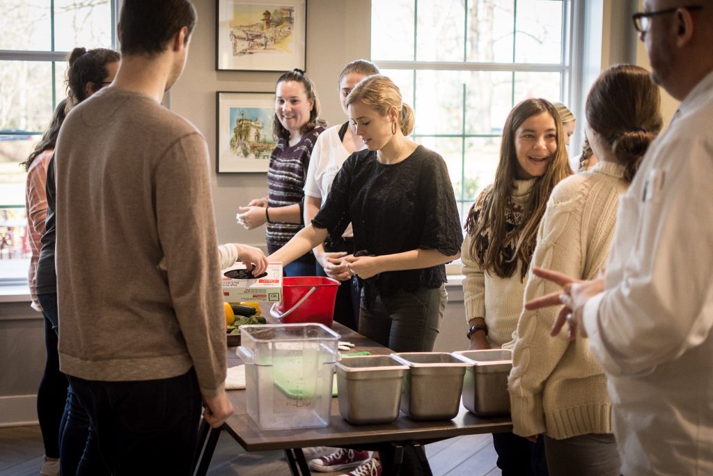 Students gather around tables that have been set up to prepare ingredients for a cooking class. The students are smiling and visibly excited about the experience.