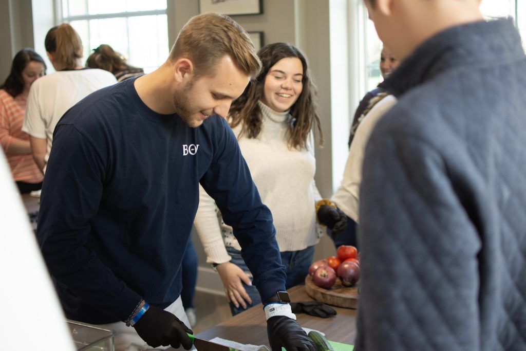 A student wearing a dark-colored long-sleeved shirt uses a kitchen knife to chop up dark green cucumbers as other students look on and smile.