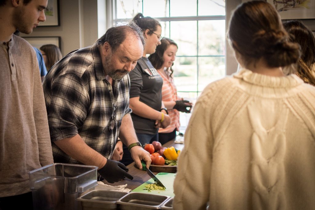 Boaz stands over some ingredients as he cuts them with a chef's knife and gloved hands.