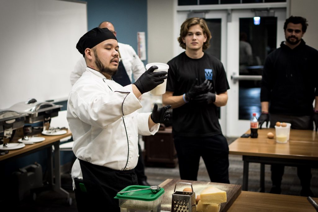 Elon Dining's Chef Danny holds up a half-full deli container of a white powder, showing it to a student dressed in all black.