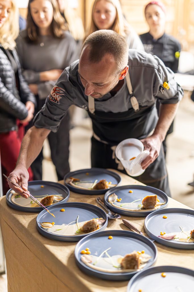 Elon dining's Chef Brandon stands over a number of gray plates as he adds garnish and finishes preparation during a teaching kitchen experience. There are numerous students in the background smiling and looking on.