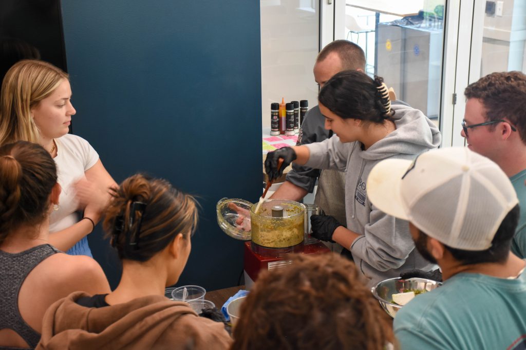 A student stirs a mixture being processed in a food-processor. Chef Brandon stands beside her, providing encouragement and guidance.