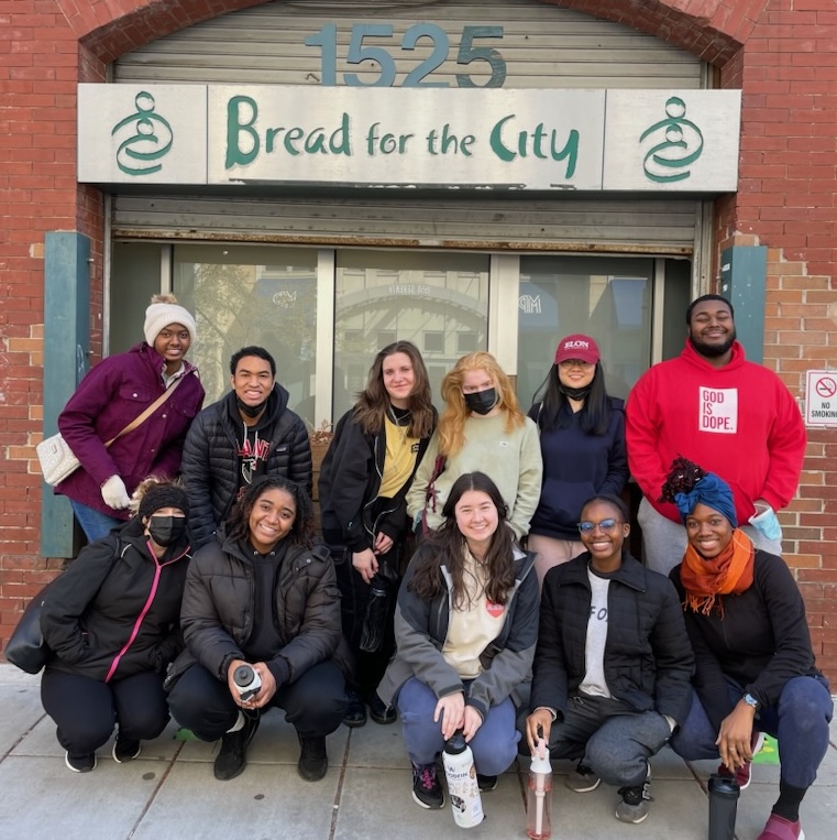 A group of students pose for a group photo underneath the sign "Bread for the City."