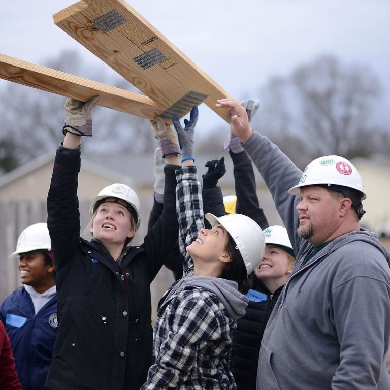 A group of students wearing hard hats lift pieces of lumber in a housing build.