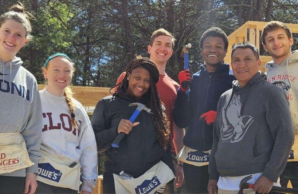 A group of Elon students and professors pose for a picture. They are wearing Lowe's work aprons and two students are holding hammers. 