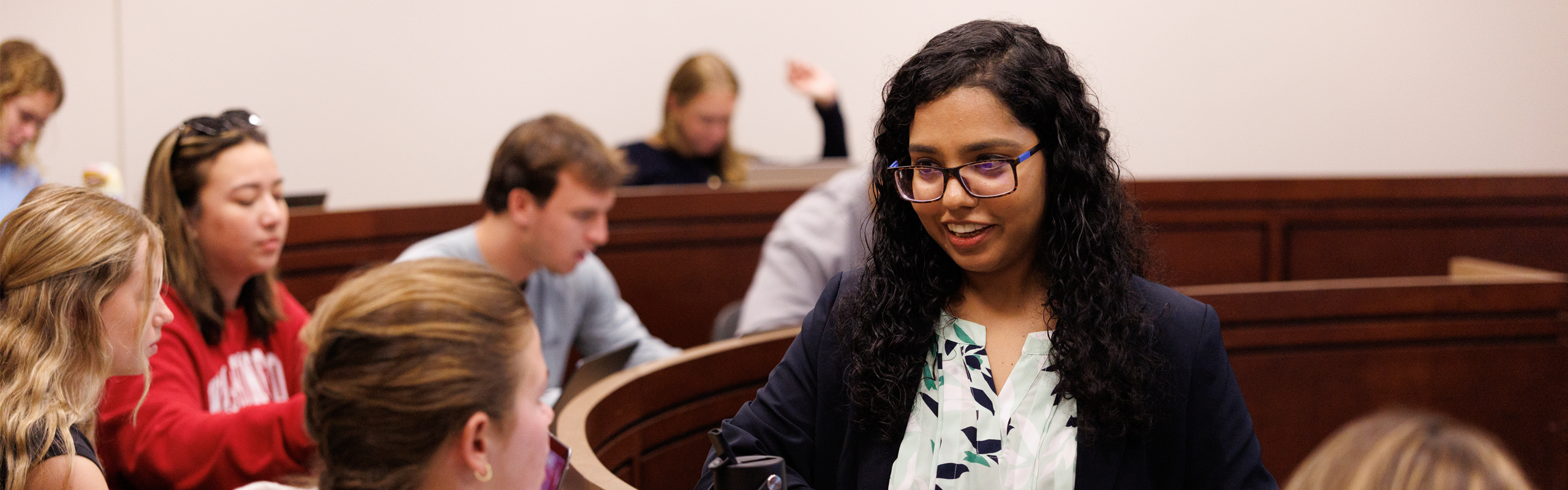 Smaraki Mohanty speaks with a student during class in a tiered lecture hall. The student gestures while seated at a laptop, and several other students in the background take notes or use computers during the discussion.