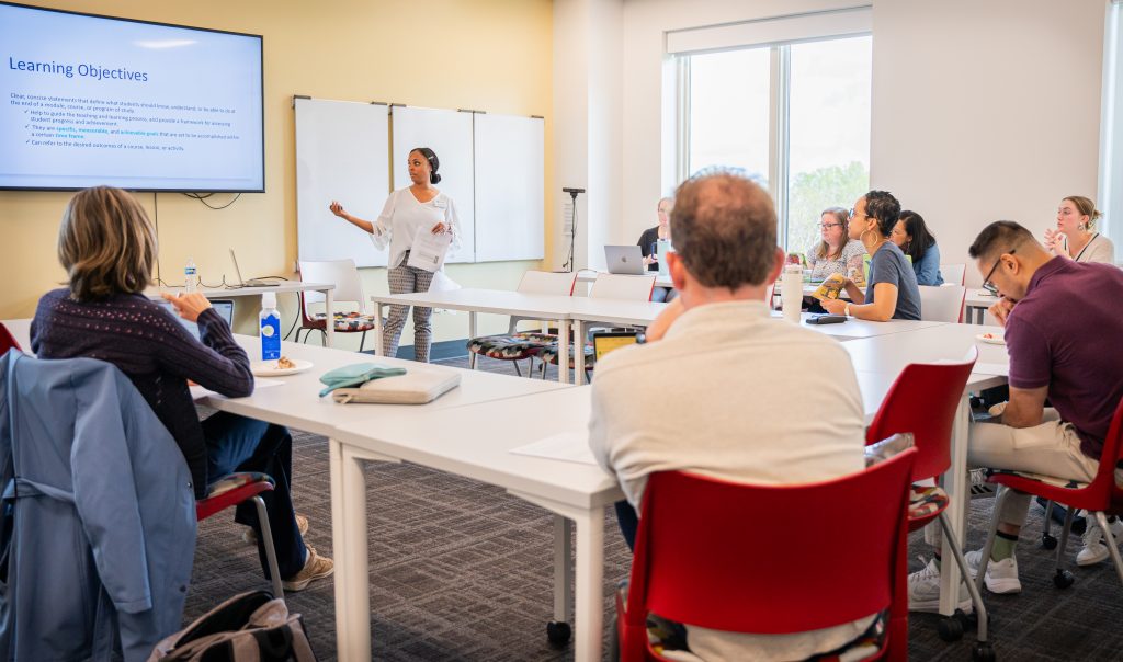 A woman stands at the front of a classroom presenting a slide titled 'Learning Objectives' to a group of faculty and staff seated at tables. Participants listen attentively, some using laptops or taking notes, in a bright modern learning space with large windows and whiteboards.