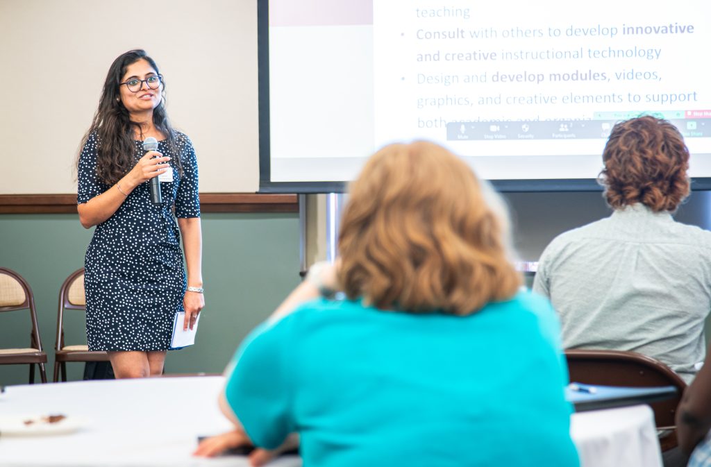 Indian woman wearing a navy dress with white polka dots and black rimmed glasses holds a microphone while giving a presentation to a room of people. She stands on the left side of the photo with the projector screen taking up the center-right two-thirds of the background. Spectators are out of focus in the foreground of the image.