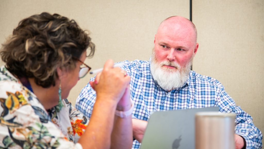 Bald white man with white beard wearing a blue paid button-down shirt is consulting a faculty member. The man sits on the right side of the photo