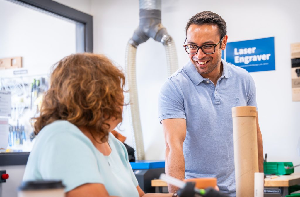 A man in a light blue polo shirt smiles and leans forward while engaging in conversation with a woman seated at a worktable in a makerspace. The room contains tools, a laser engraver sign, and various project materials, suggesting a creative, hands-on environment.