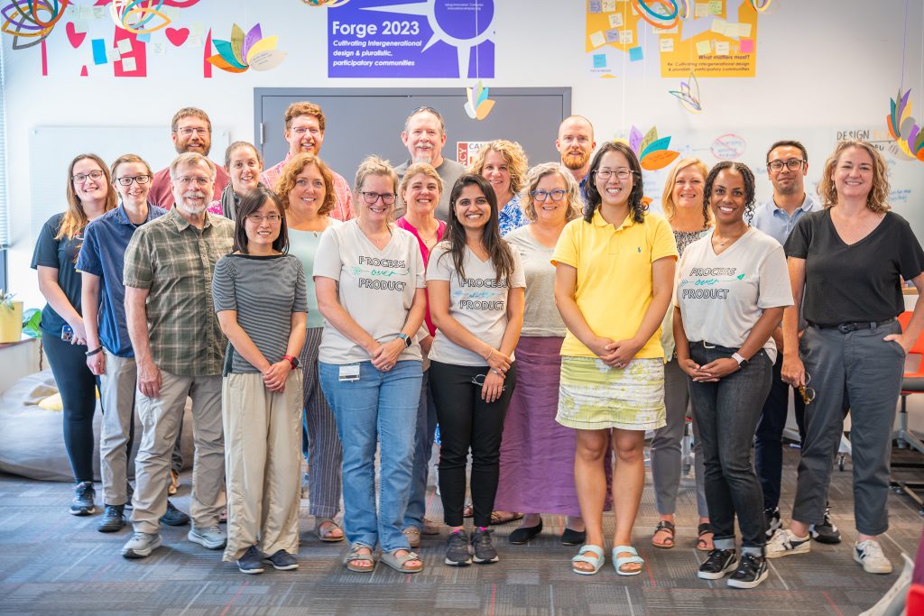 A group of smiling faculty and staff stand together in a brightly decorated room at Elon University for a group photo during Forge 2023. Colorful paper decorations and posters with design thinking themes, including one that reads 'Process Over Product,' are visible on the walls behind them. The setting conveys a welcoming, creative, and collaborative atmosphere