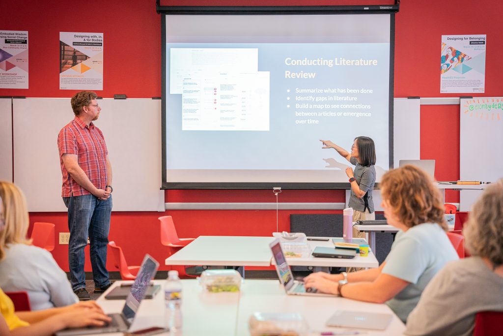 A woman gestures toward a large projection screen while leading a session on 'Conducting Literature Review' in a classroom with a red accent wall. The slide includes bullet points about summarizing research, identifying gaps, and mapping connections between articles. A man stands to the side near a whiteboard, and several seated participants with laptops listen and take notes. Posters about design thinking and community engagement hang on the walls.
