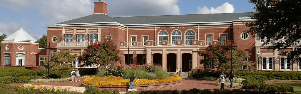 Wide exterior view of Moseley Center at Elon University, a large red-brick campus building surrounded by landscaped gardens and blooming flowers. Students walk along brick pathways in front of the building under a partly cloudy sky.