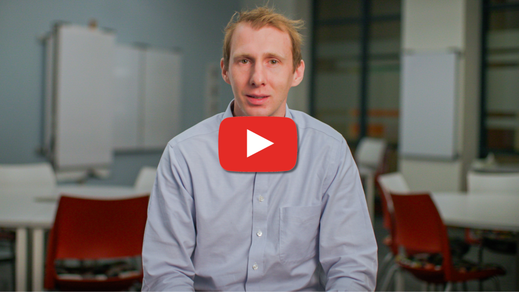 A man in a light blue button-down shirt sits facing the camera in a classroom or office setting, with a large red play button overlay centered on the image indicating a video. Tables, chairs, and whiteboards are visible in the softly blurred background.