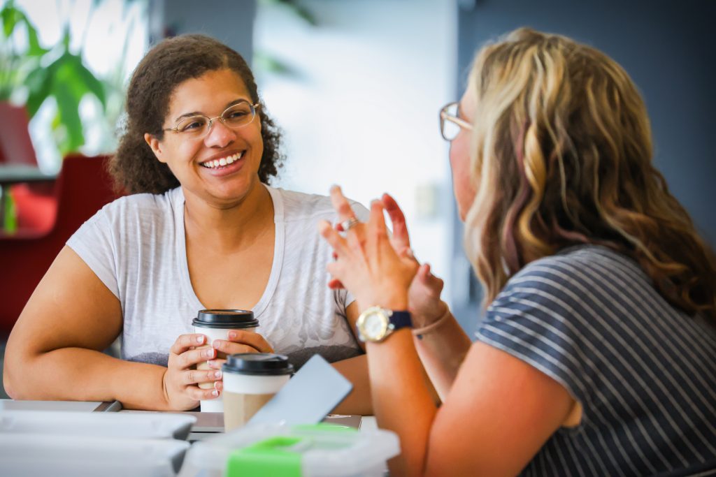 Two women sit across from each other at a table, smiling and engaged in conversation while holding coffee cups. One woman gestures with her hands as she speaks, creating a warm and collaborative atmosphere in a casual campus or office setting.