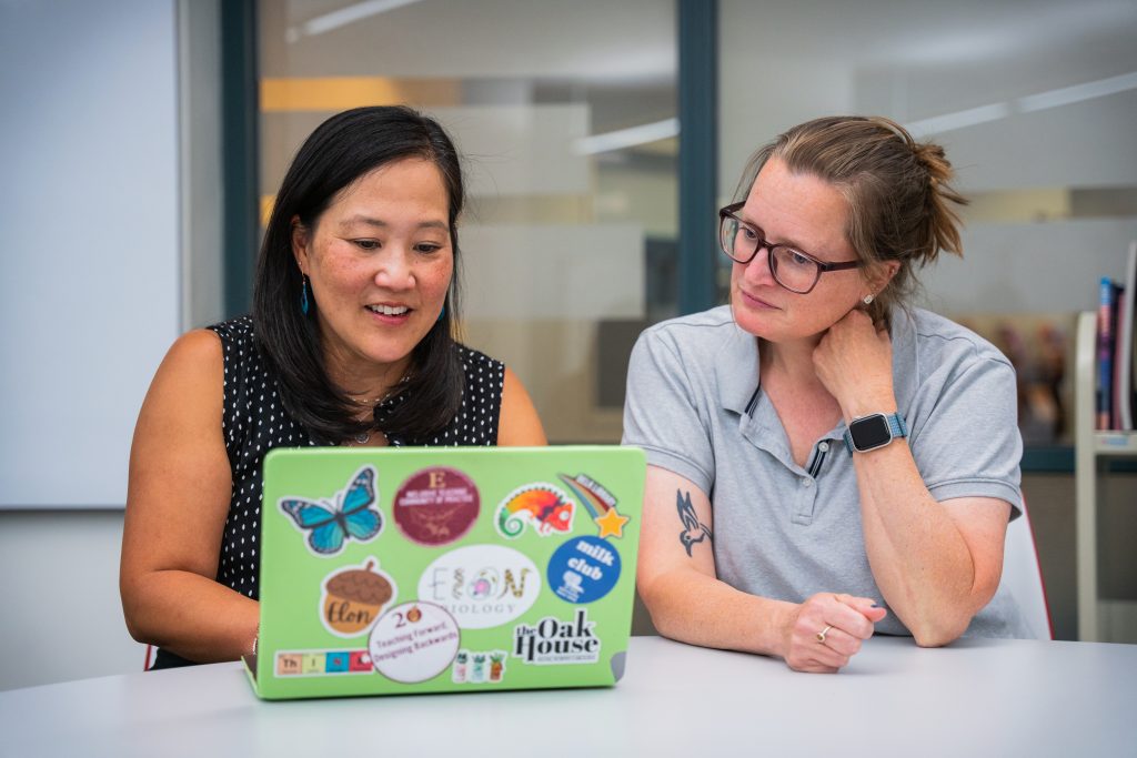 Two women sit side by side at a table, collaborating while looking at a laptop covered in colorful stickers, including Elon-themed designs. One woman gestures as she speaks, while the other listens attentively with her hand near her neck. The setting appears to be a modern office or classroom space with glass walls in the background.