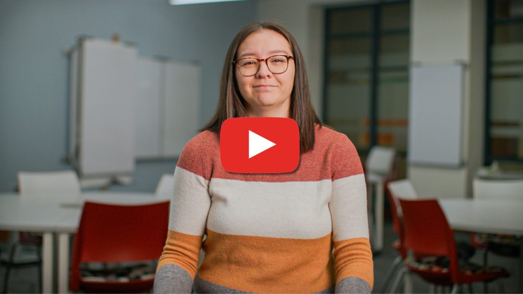 A woman wearing glasses and a color-block sweater stands facing the camera in a classroom setting, with a large red play button overlay indicating a video. Tables, chairs, and whiteboards are visible in the background.