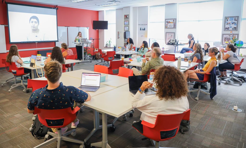 Faculty and staff attend a Teaching and Learning Technologies workshop, seated at collaborative tables while viewing a presentation and video on a large screen. Participants engage with laptops and discussion materials in a bright, technology-rich learning environment.