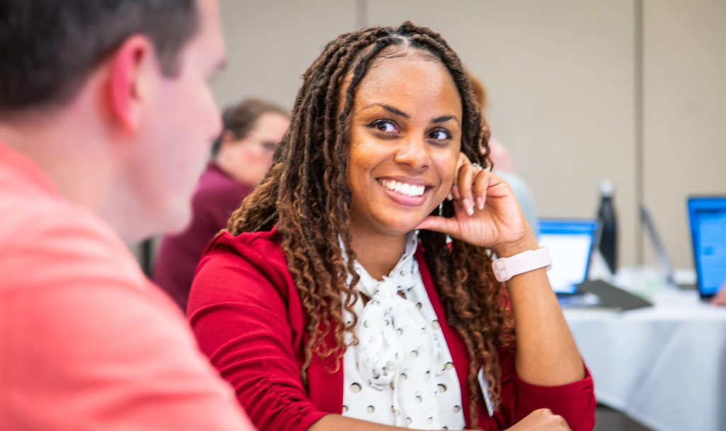 A woman in a red blazer and patterned blouse smiles while engaged in conversation during a meeting or workshop. Seated at a table with colleagues, she rests her head on one hand and looks attentively toward another participant, while laptops and attendees are visible in the softly blurred background.