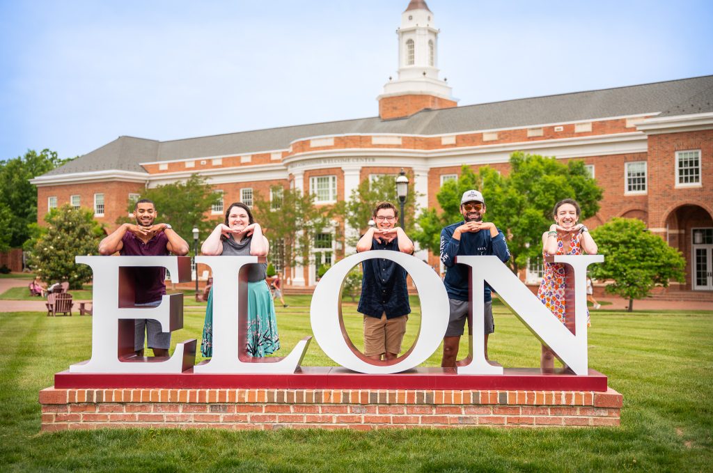 Five smiling students pose behind the large white ELON sign on the lawn in front of the Inman Admissions Welcome Center at 51������. Each student rests their chin on their hands in a playful pose, with the red-brick building and cupola visible in the background on a sunny day.