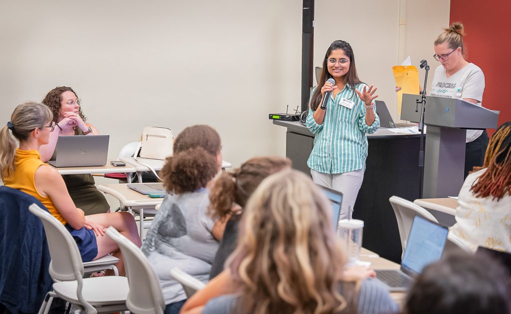 A woman in a green-and-white striped shirt speaks into a microphone while addressing a group during a workshop or professional development session. Participants seated around the room listen attentively, some using laptops, while another facilitator stands at a podium reviewing notes. The setting is a collaborative classroom environment focused on discussion and learning.