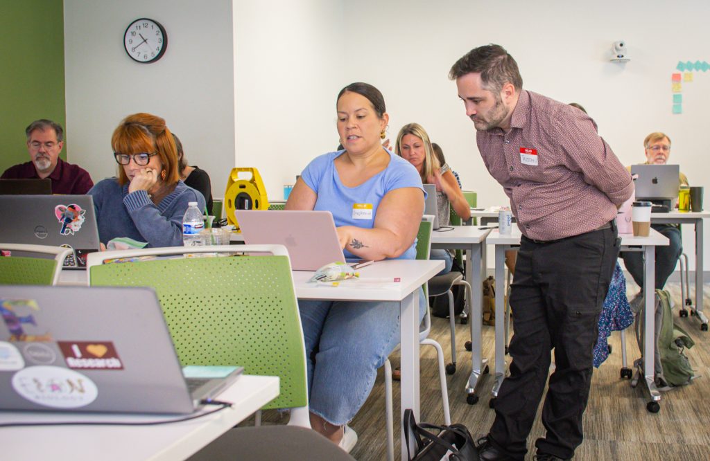 A facilitator provides individualized support to a participant using a laptop during a technology workshop. Other faculty and staff are seated throughout the classroom, actively working on laptops and participating in the hands-on learning session.