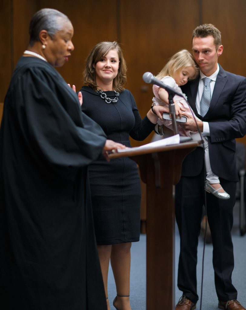 Carrie Vickery takes an oath, raising her right hand. Her family is nearby. Another judge is administering the oath.