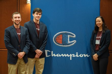 Three students standing by a Champion sign
