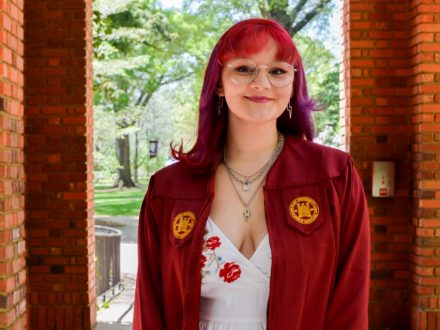 Smiling woman with red hair wearing glasses, a white dress and maroon graduation gown standing outdoors.