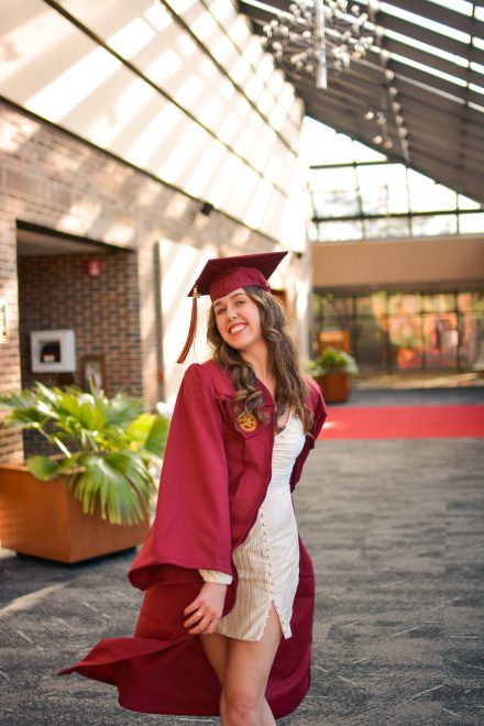 Picture of a woman in a cap and gown smiling in a theater lobby.