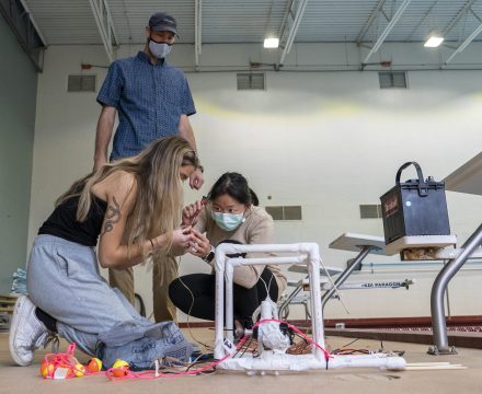 Two students connect parts to a square frame made of PVC pipe, a prototype underwater remotely operated vehicle, as a professor watches.